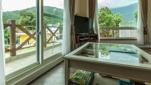 Una habitación con balcón, televisor sobre un mueble y una mesa de vidrio con revistas.