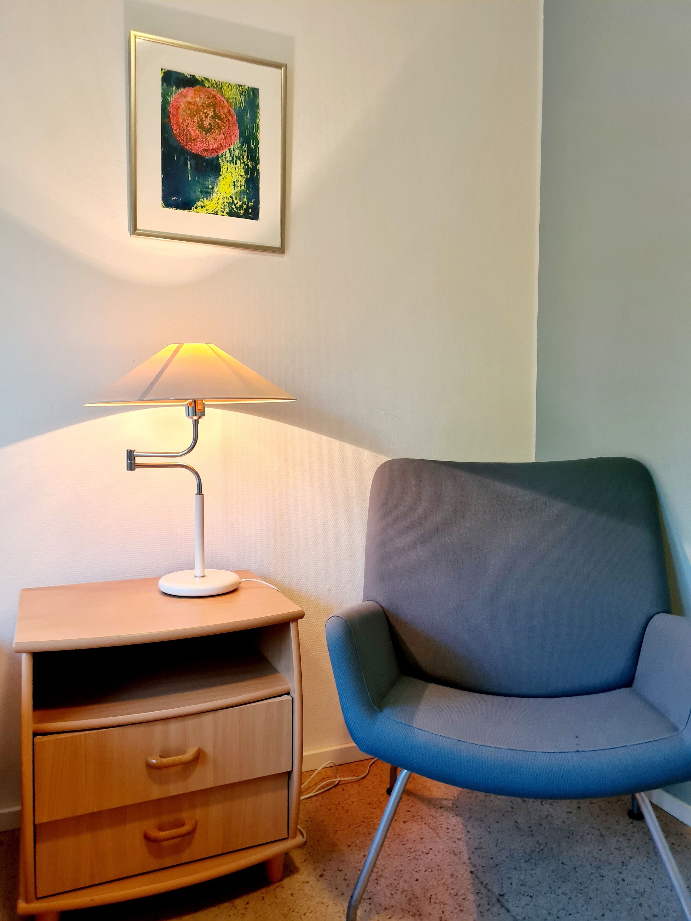 A room with a wooden nightstand, a lamp, a blue chair, and a framed abstract painting on the wall.