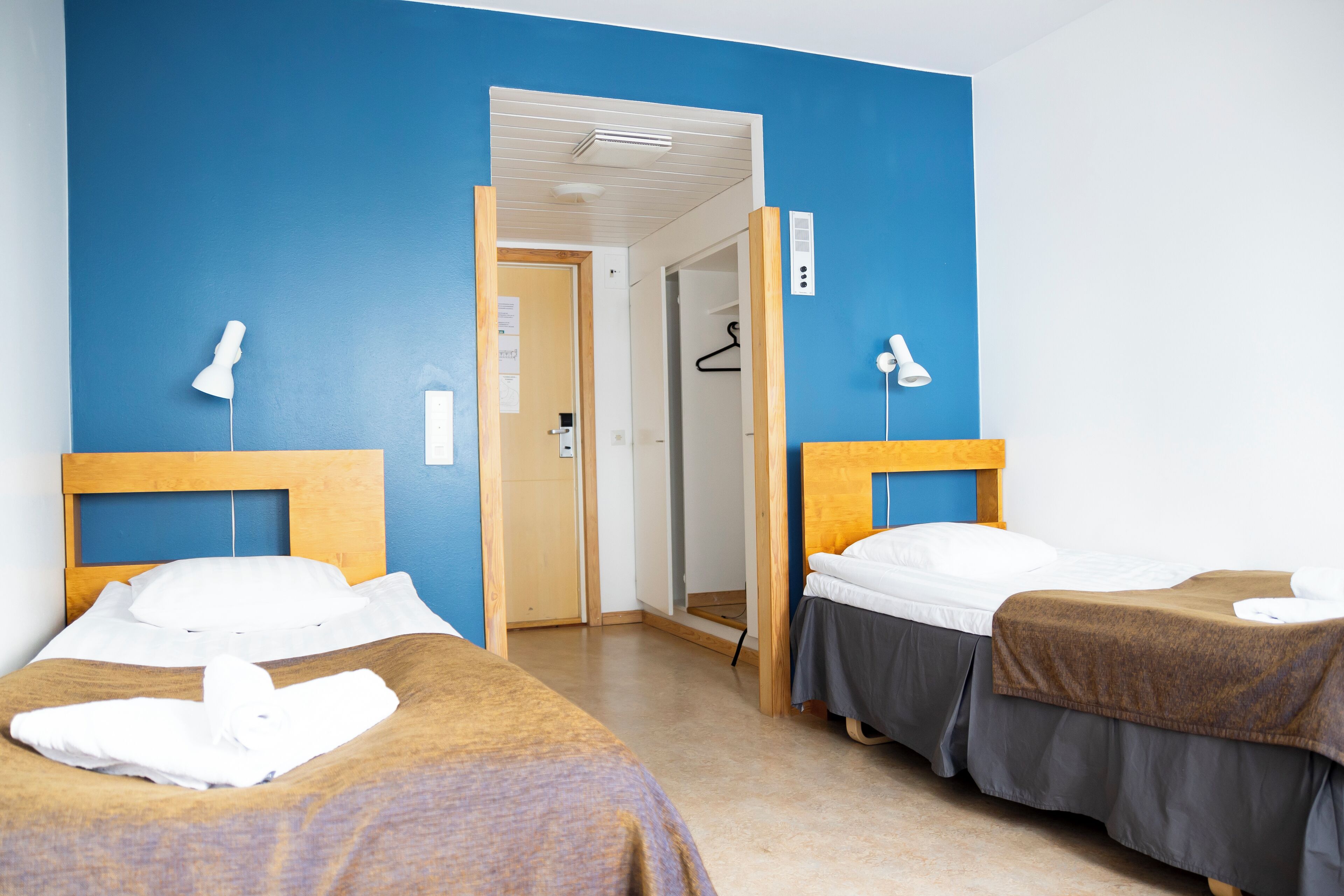 Hotellihuone, jossa on kaksi sänkyä, puinen pääty ja sininen seinä. Ovi johtaa kylpyhuoneeseen.