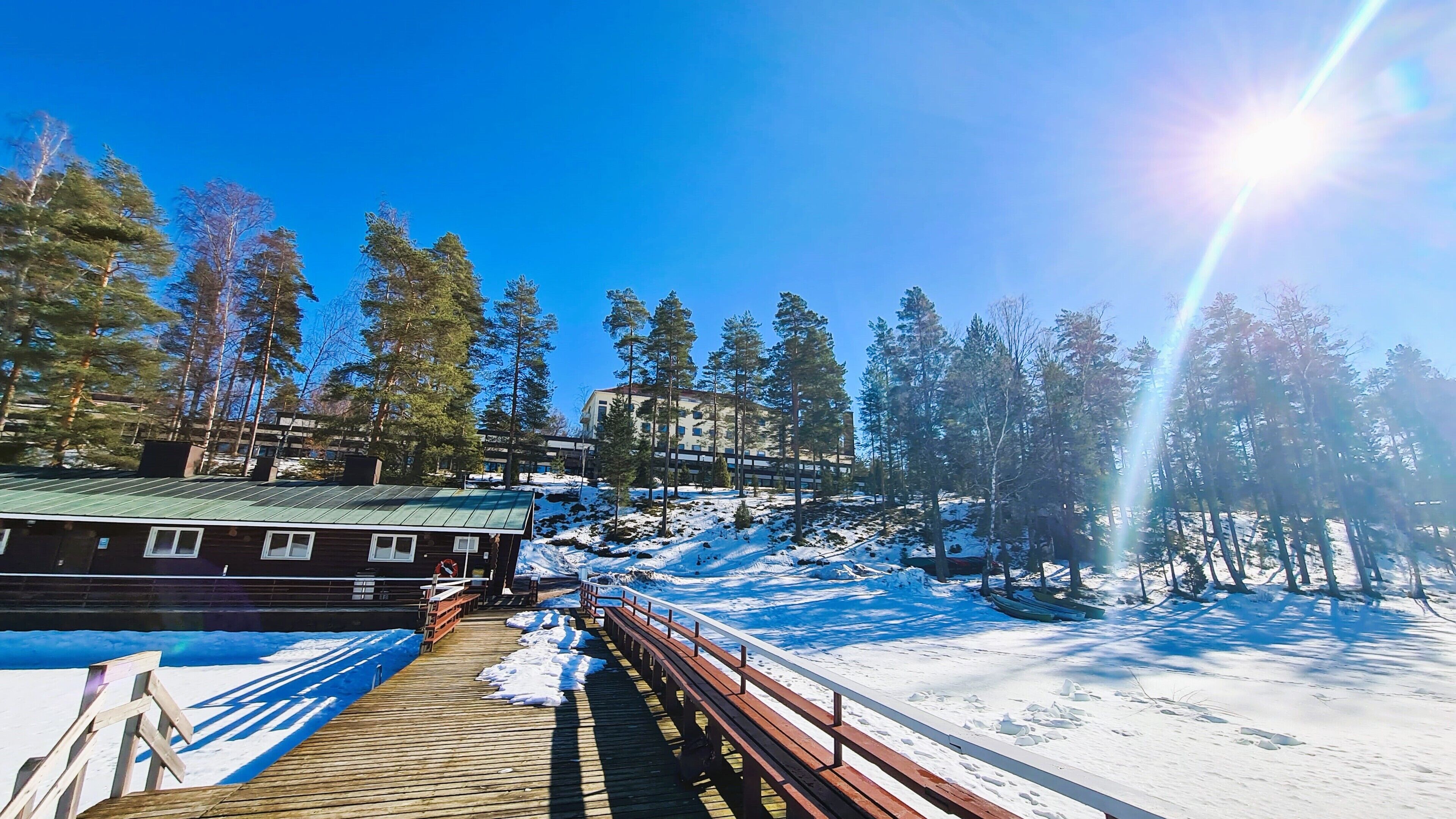 Una terraza de madera que conduce a un edificio con techo verde, rodeado de nieve y árboles.