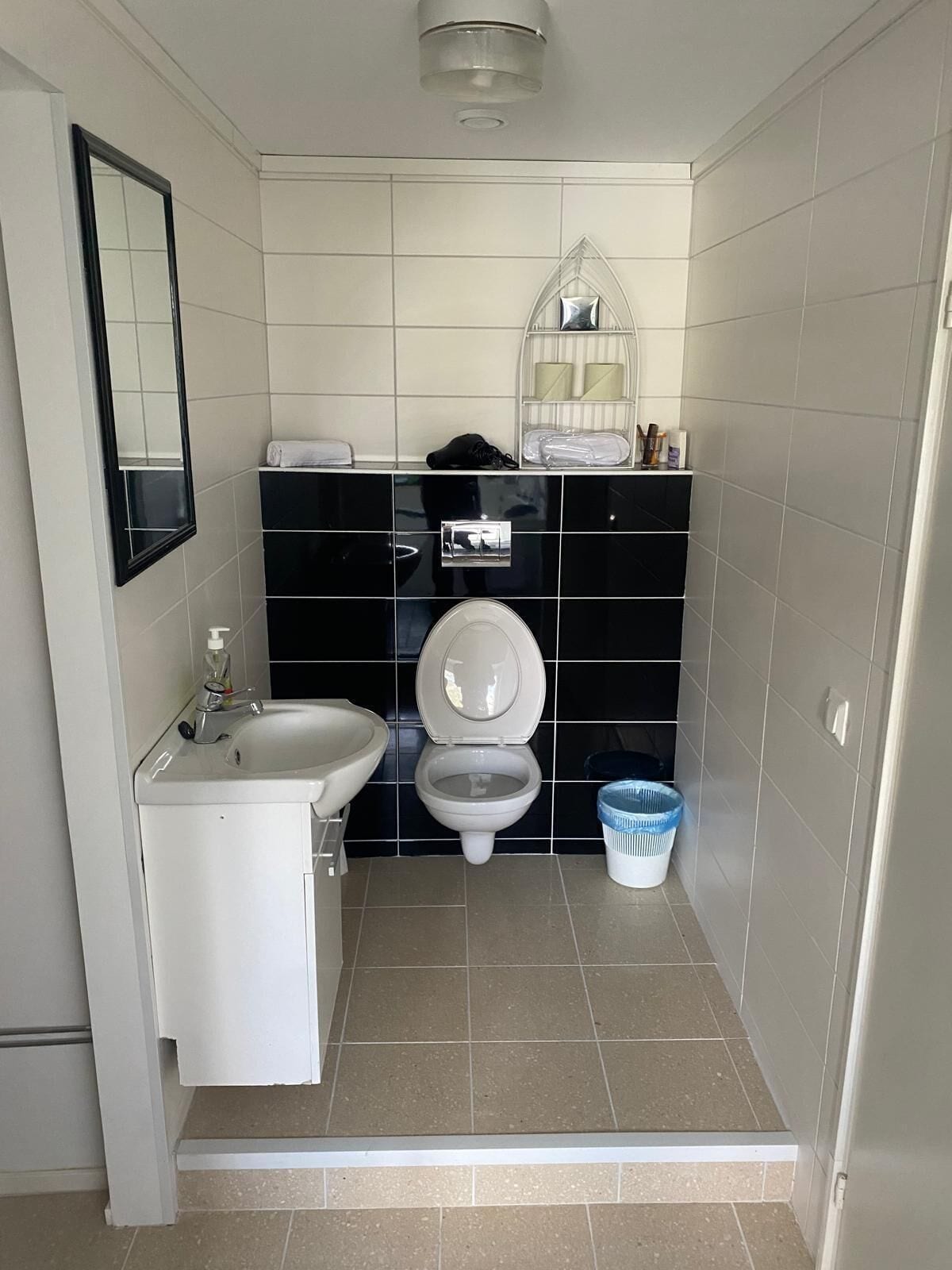 A compact bathroom with a white sink, black tiled walls, a toilet, and a blue laundry basket.