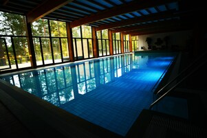 Una piscina interior con paredes de vidrio transparente, vigas de madera y suelo de baldosas.