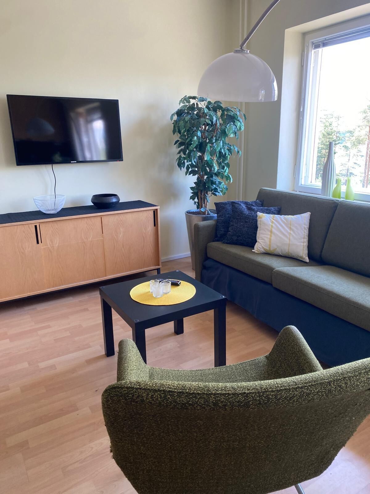 A modern living room with a flat-screen TV, a wooden cabinet, a black coffee table with a yellow placemat, a grey sofa with blue and white cushions, a green armchair, a large potted plant, and a white pendant lamp.