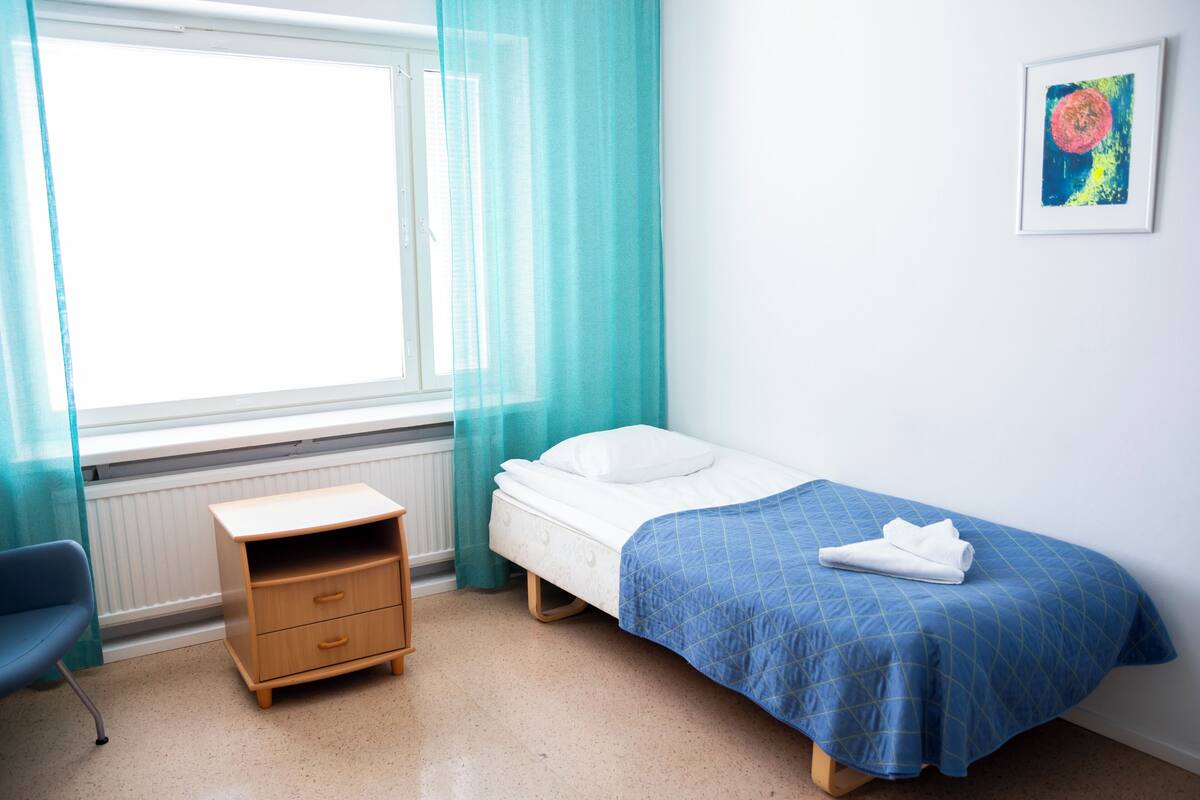 A bedroom with a bed, a nightstand, a chair, a window with sheer curtains, and a framed picture on the wall.