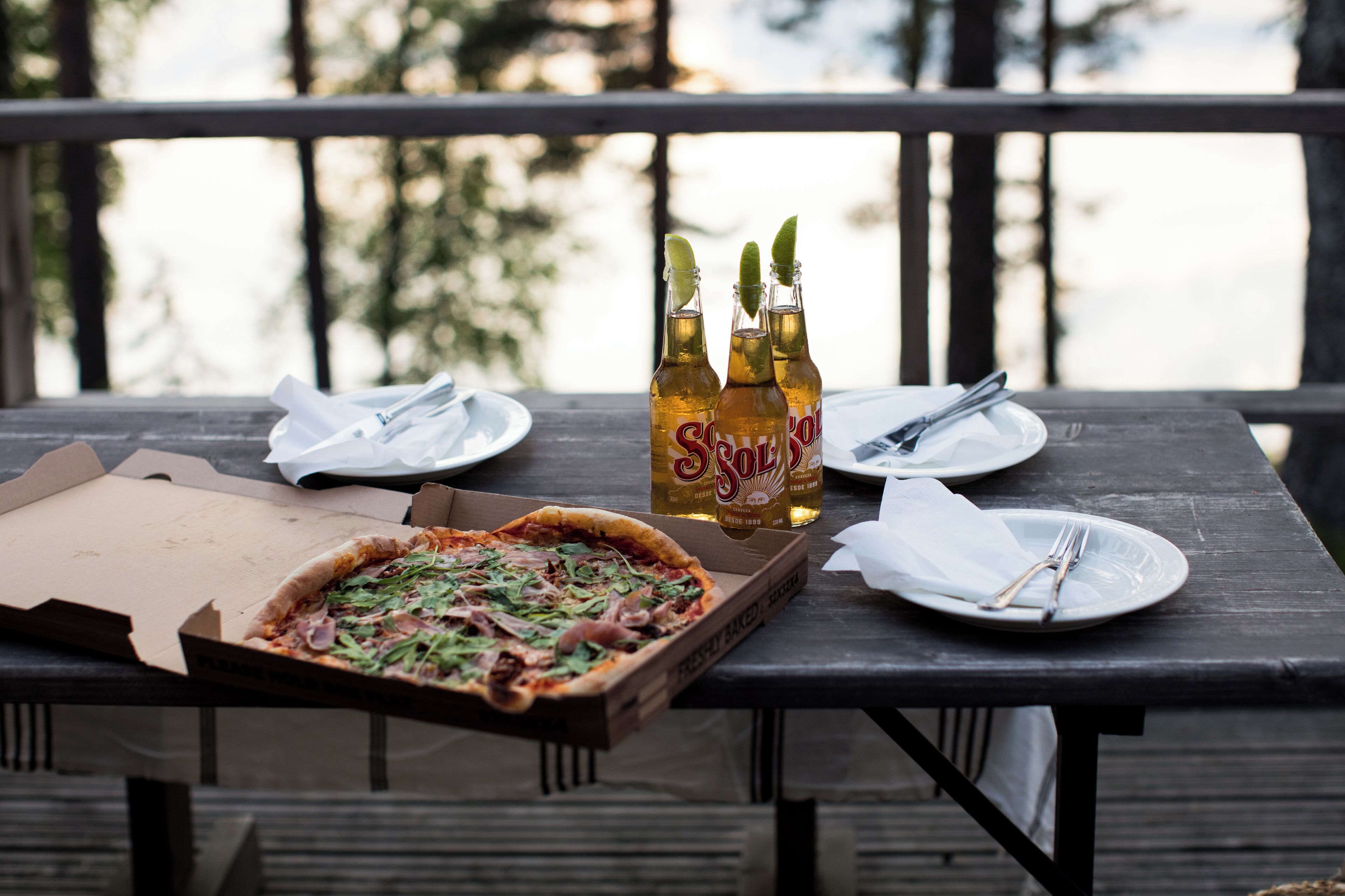 A wooden table with a pizza box, two bottles of Sol y Sol beer, and plates with cutlery on a balcony.