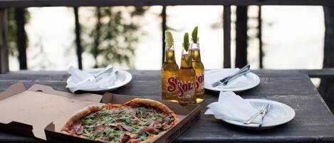 A wooden table with a pizza box, two bottles of Sol y Sol beer, and plates with cutlery on a balcony.