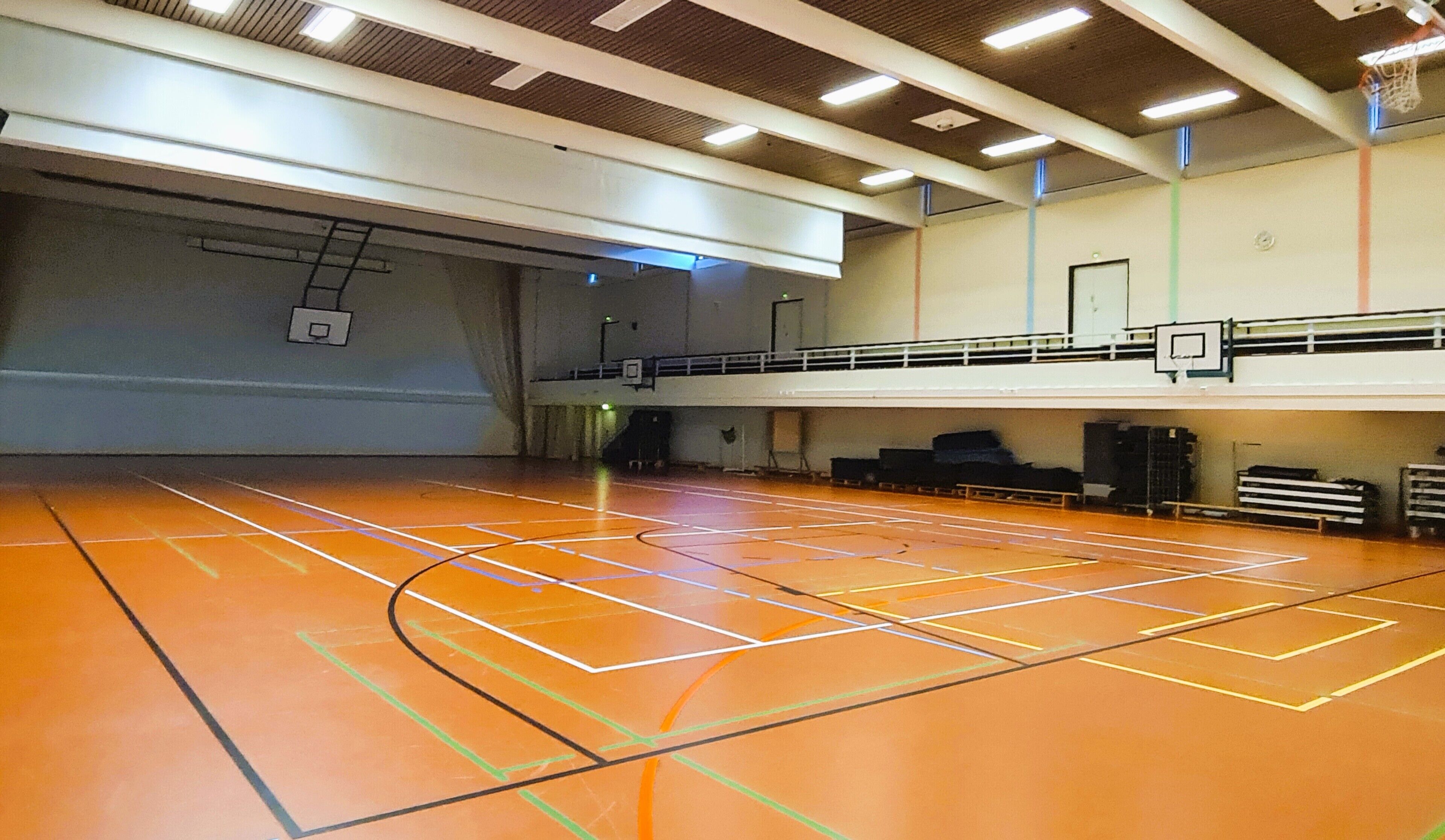 An indoor basketball court with a wooden floor, a basketball hoop, and a mezzanine level with seating.