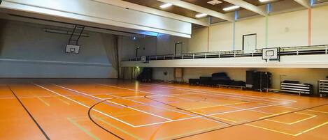 An indoor basketball court with a wooden floor, a basketball hoop, and a mezzanine level with seating.