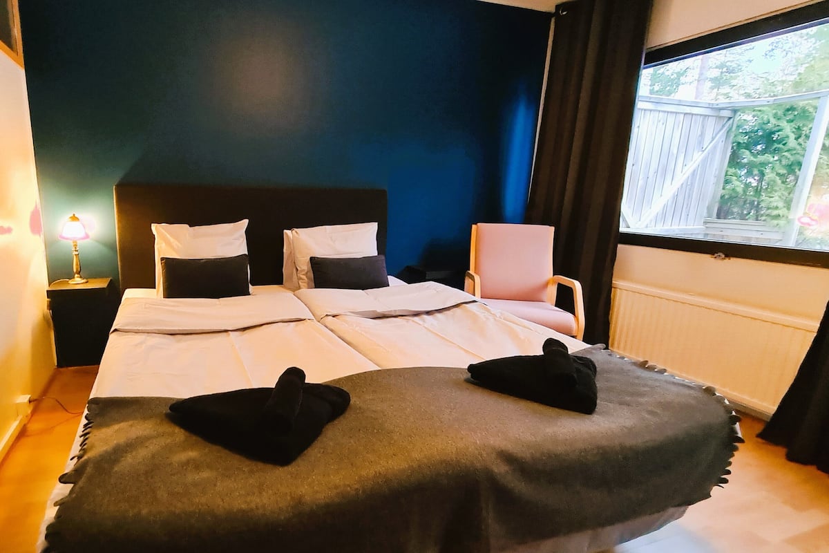 A neatly made bed with white linens and two black pillows, a bedside lamp, a chair, and a window with a view of greenery.