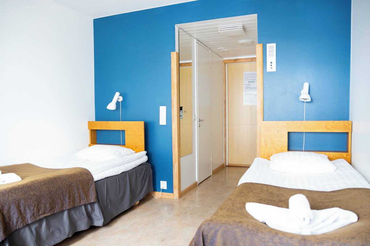A hotel room with two single beds, each with a wooden headboard, white bedding, and a brown throw blanket. There is a wooden door leading to a bathroom, and a white wall-mounted lamp on each bed.