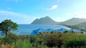 Ein Küstenhotel mit mehreren Gebäuden, freiem Blick auf das Meer und einem markanten Berg im Hintergrund.