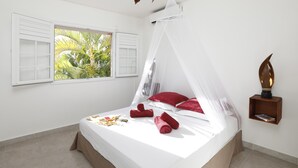 Ein Schlafzimmer mit einem Himmelsbett, einem Fenster mit Fensterläden, einem Nachttisch mit Lampe und Blick auf Palmen im Freien.