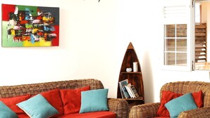 Ein Wohnzimmer mit einem roten Sofa, blauen Kissen, Korbstühlen, einem Couchtisch mit Büchern und einem Hut sowie einem Bücherregal mit Zeitschriften.