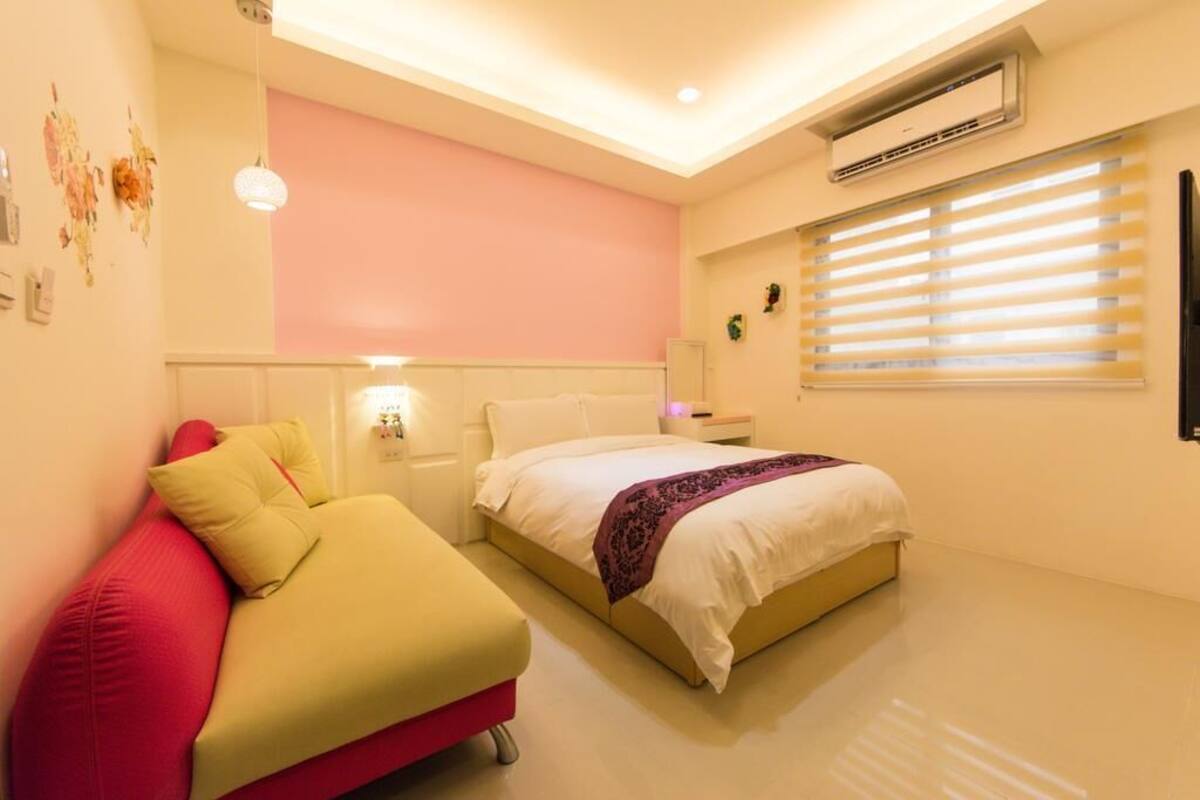 A bedroom with a bed, a red couch, a bedside table, and an air conditioning unit.