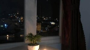 Una habitación con vista a la ciudad de noche, una mesita con una planta en maceta y dos velas, y un ventanal con cortinas.
