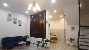 Amplia sala de estar con un sofá azul oscuro, una mesa de centro y dos plantas en macetas. Se observa un comedor con una mesa y sillas de madera, y una cocina con estufa y armarios.