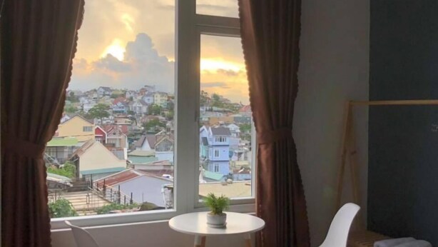 Una habitación con un ventanal grande, una mesita redonda pequeña, dos sillas blancas, una planta en maceta y un estante de madera con toallas.