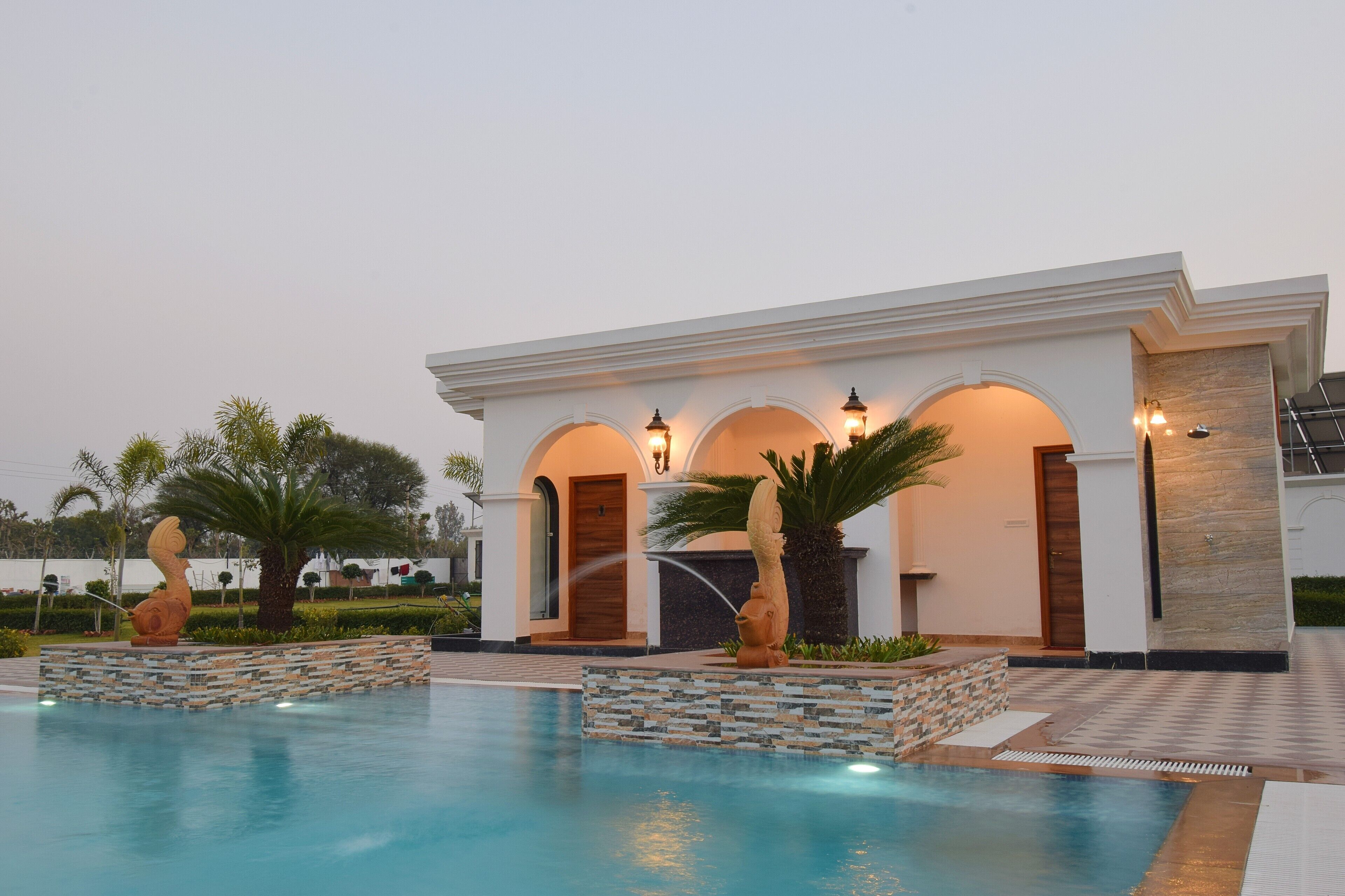 A swimming pool with a stone border and two large sculptures, a building with arched doorways and a stone facade, and palm trees in the background.