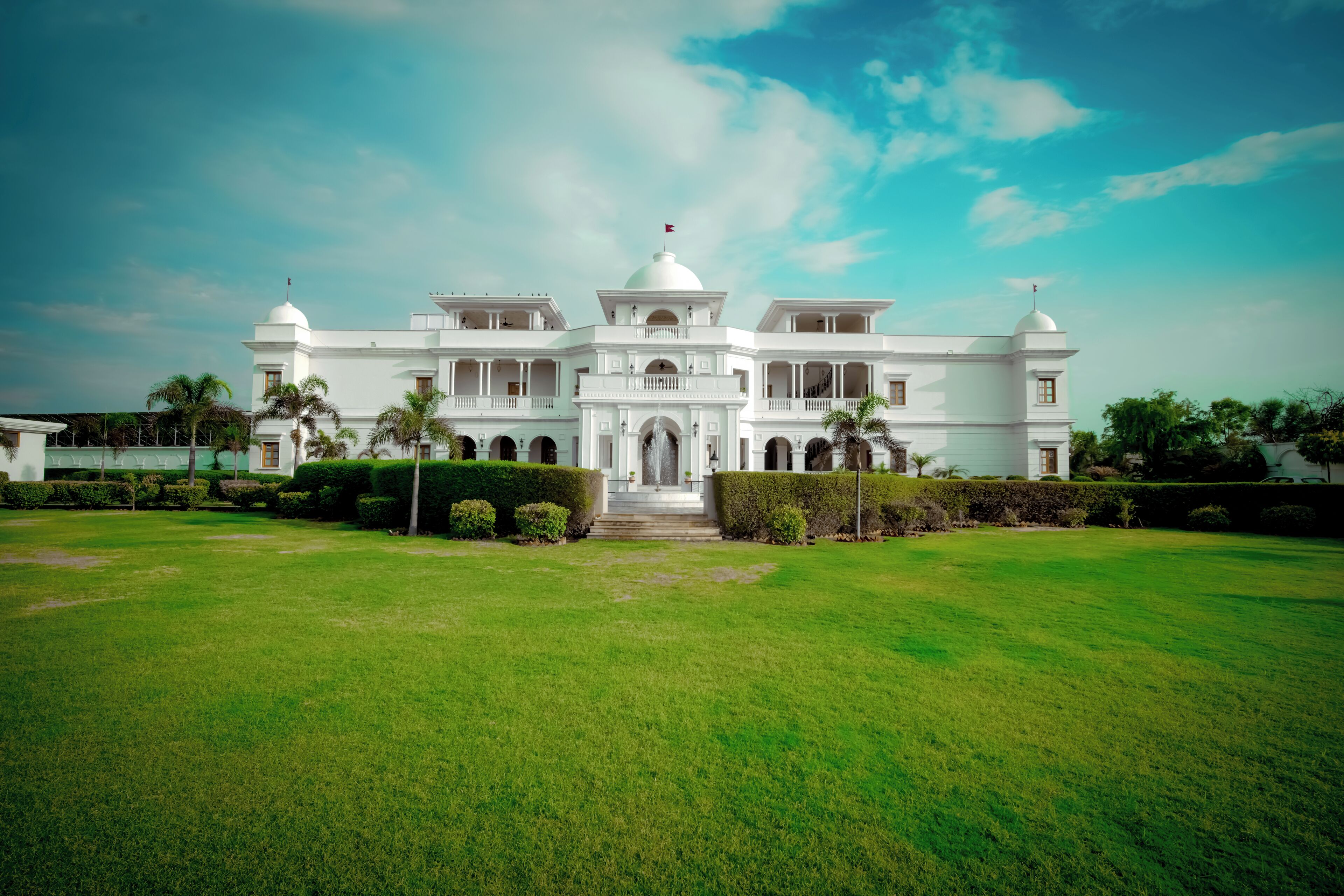A large, white colonial-style building with multiple domes and arches, surrounded by well-maintained lawns and palm trees.
