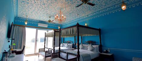 A four-poster bed with a canopy, a sitting chair, a small table, and a ceiling fan in a room with blue walls and a floral ceiling design.
