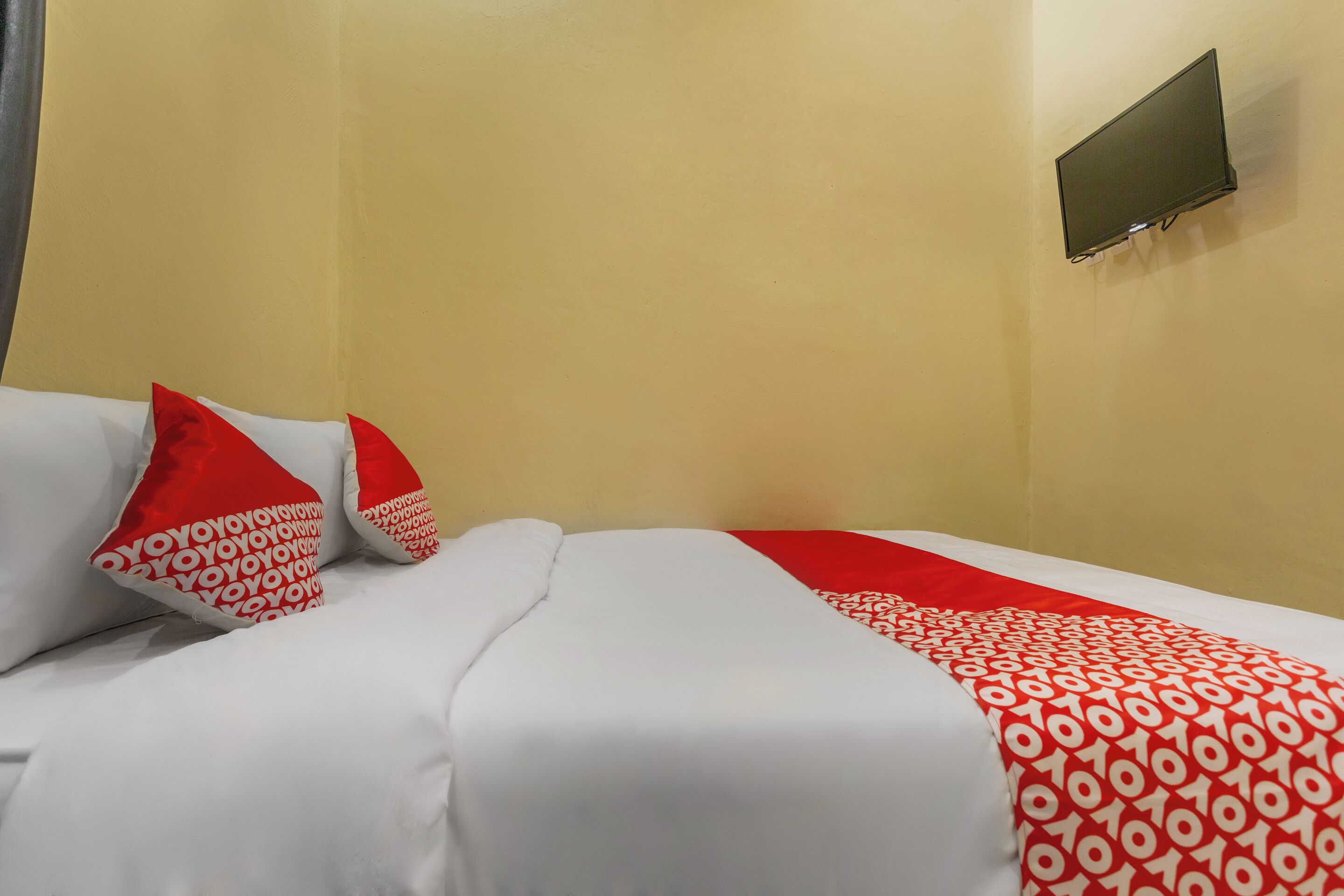A hotel room with a bed, white bedding, red and white patterned pillows, and a mounted TV on the wall.