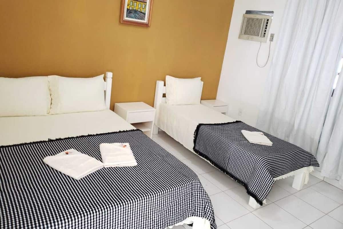 Quarto de hotel com duas camas, uma mesa de cabeceira pequena, ar condicionado fixado na parede e uma janela com cortinas brancas.
