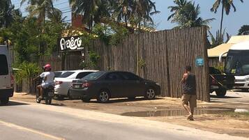 Cena de rua com cerca de bambu, carros estacionados e placa indicando um local chamado 'Pra'ero'.