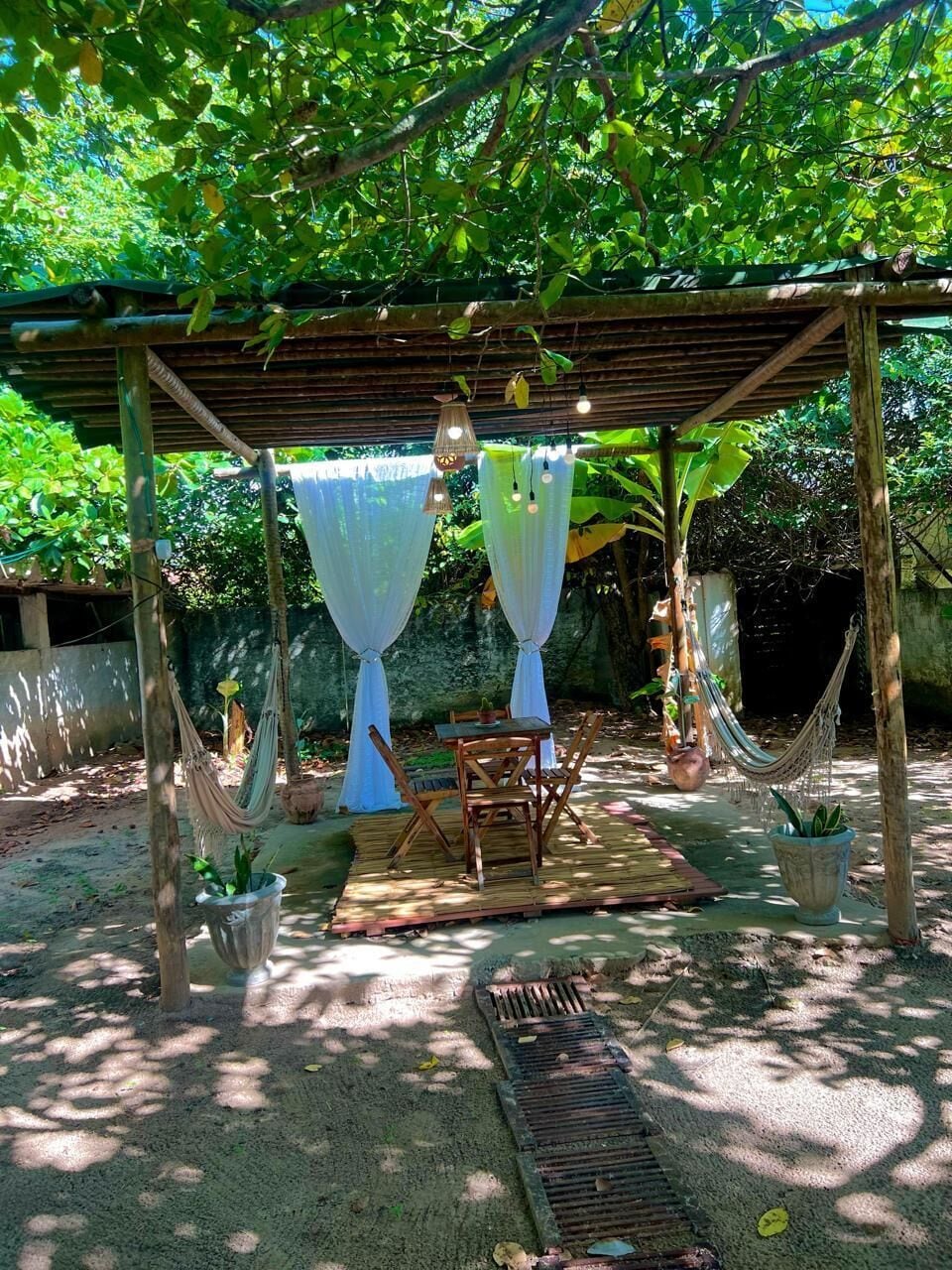 Un área de estar al aire libre techada, con una mesa y sillas, rodeada de plantas en macetas y una hamaca.