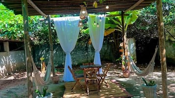 Un área de estar al aire libre techada, con una mesa y sillas, rodeada de plantas en macetas y una hamaca.
