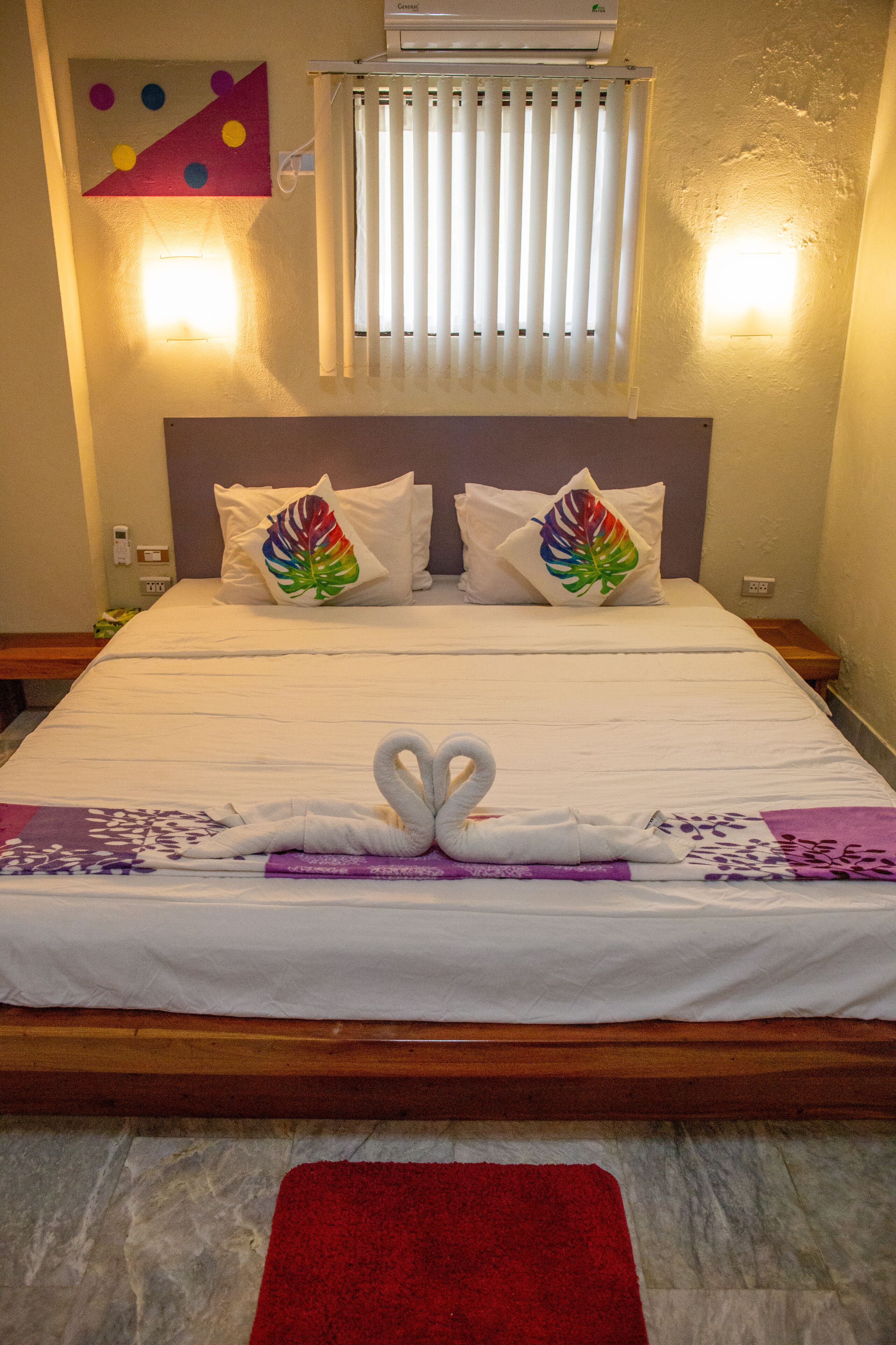 Un lit bien fait, avec du linge de lit blanc et deux oreillers ornés de motifs colorés représentant des feuilles. Un tapis rouge est placé au pied du lit.