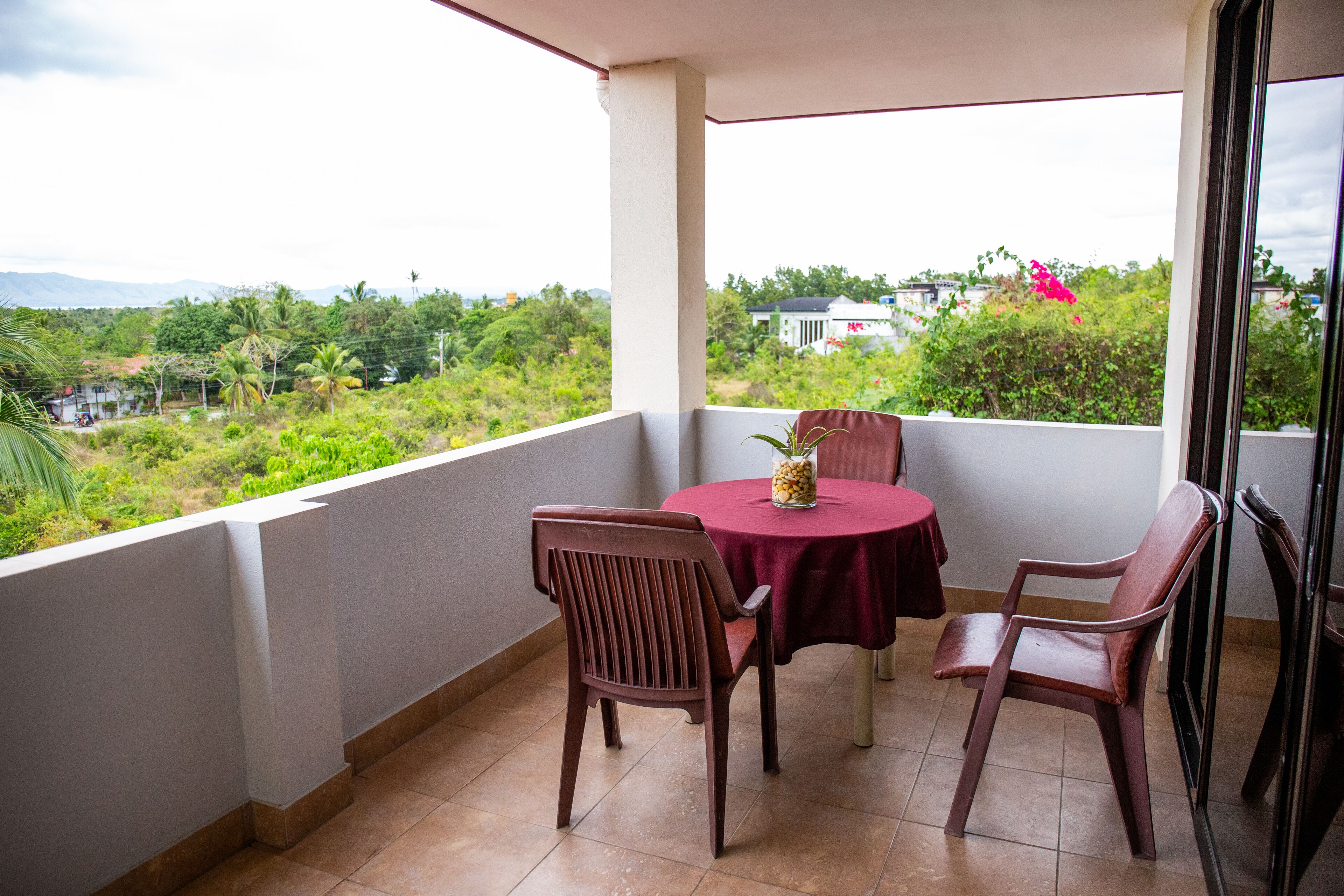 A balcony with two wooden chairs and a small table, overlooking a lush green landscape with distant buildings.
