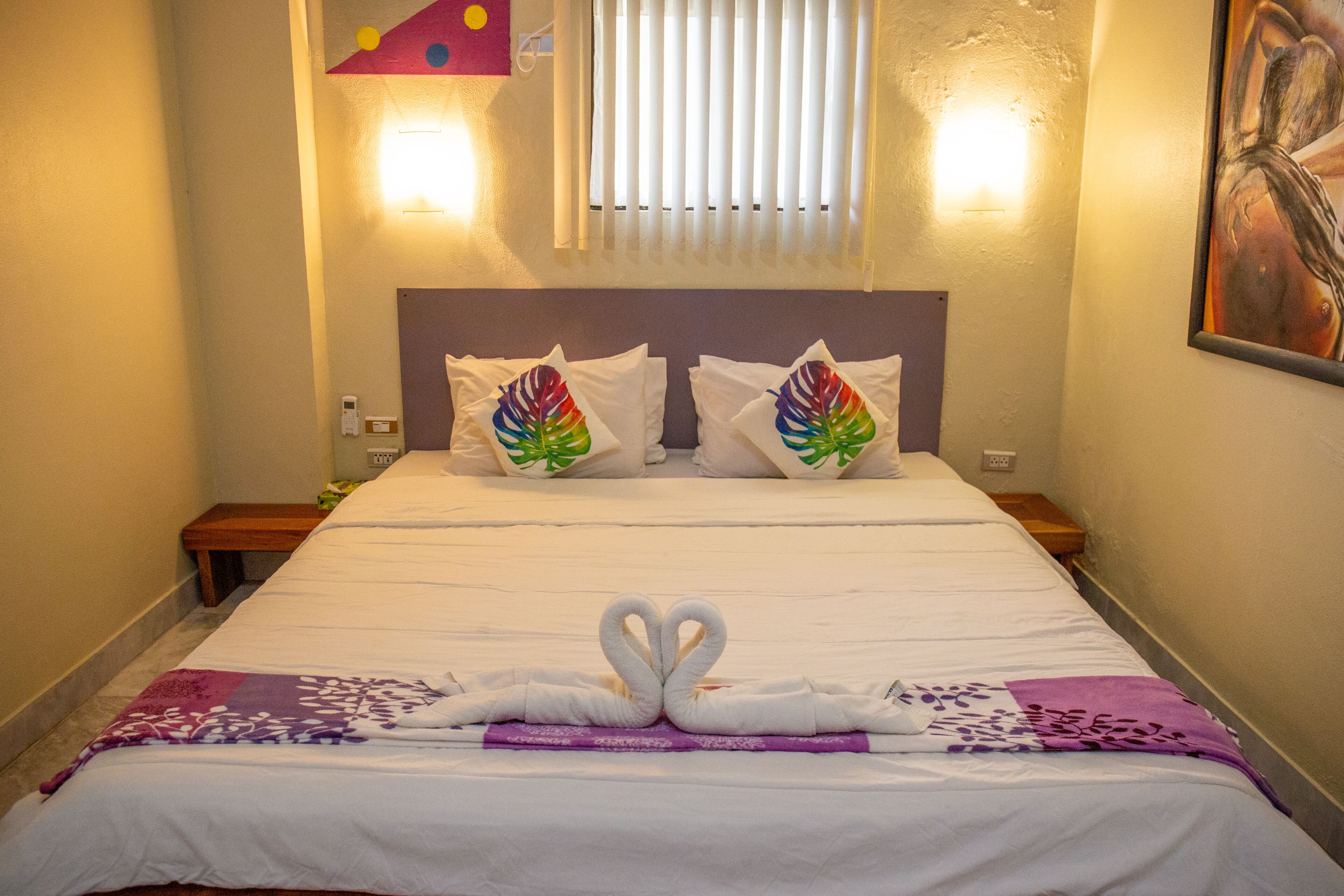 A hotel room with a bed, pillows with a colorful design, a wall-mounted light, and a painting.