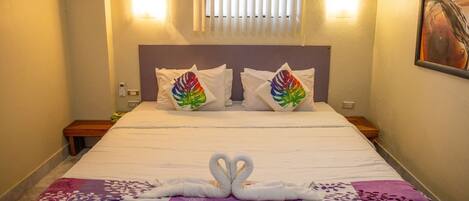 A hotel room with a bed, pillows with a colorful design, a wall-mounted light, and a painting.