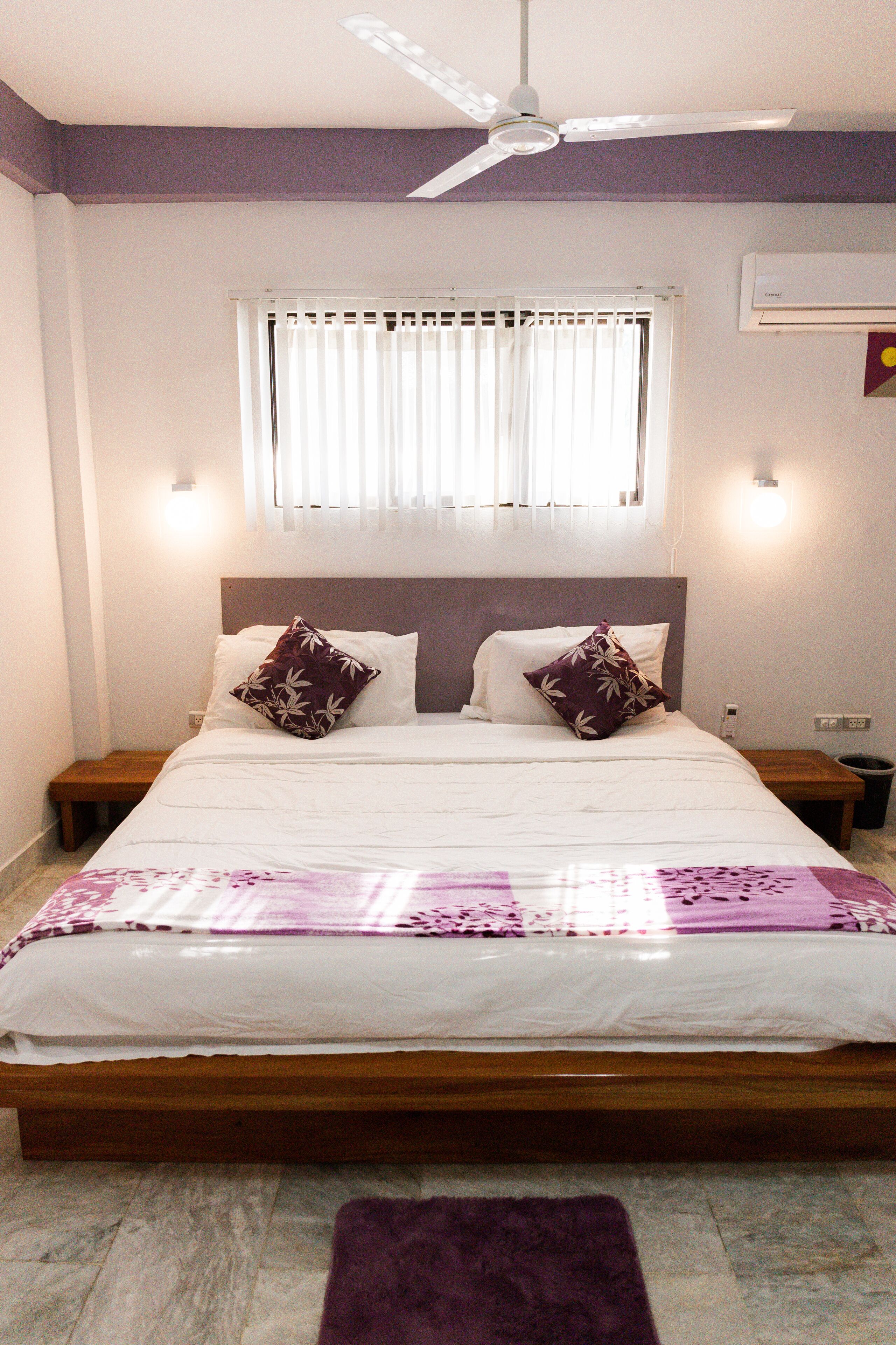 Un dormitorio con una cama, ventilador de techo, ventana con persianas y aire acondicionado.