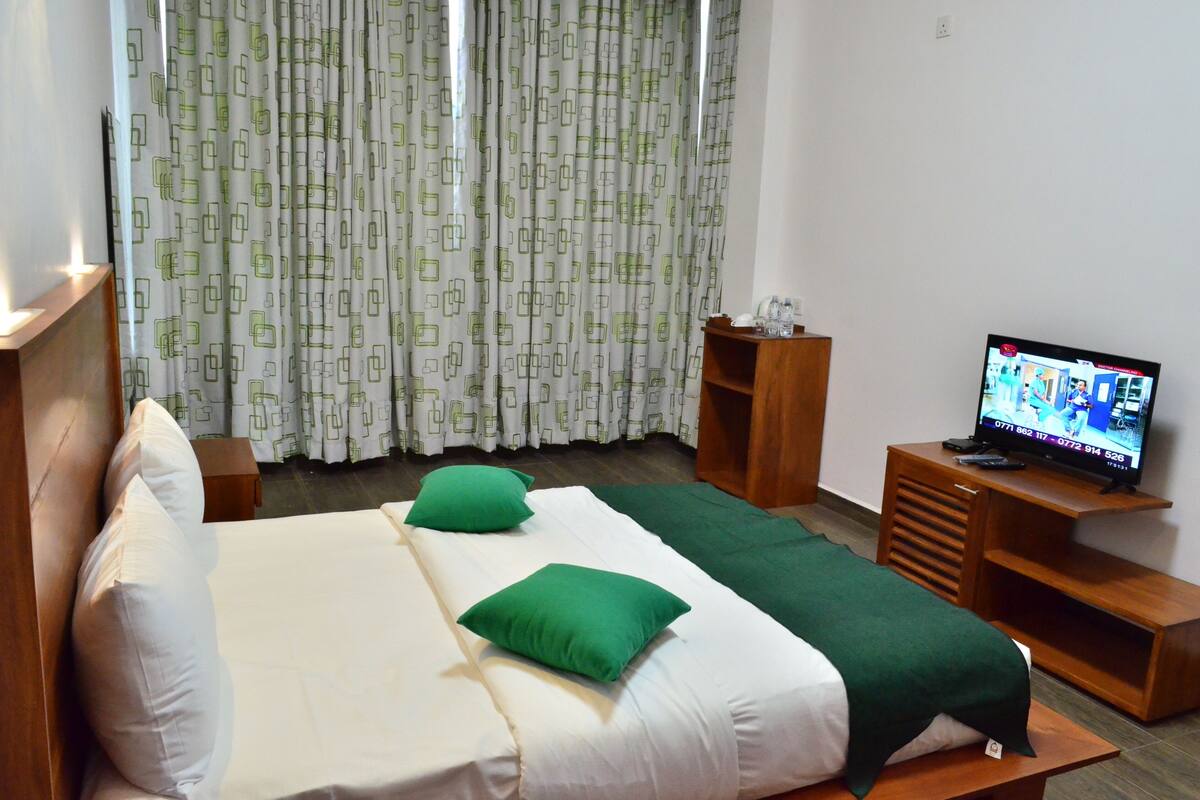 A hotel room with a bed, green pillows, a TV on a wooden stand, and curtains with a geometric pattern.