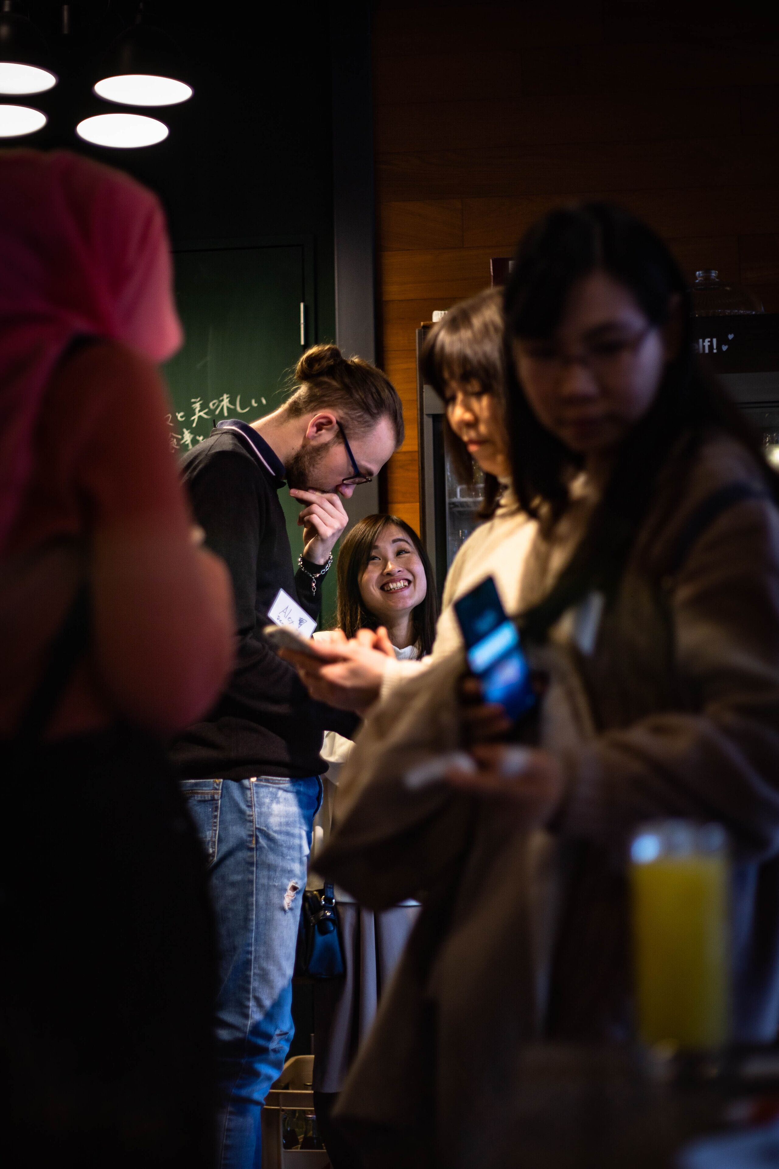 A group of people in a dimly lit indoor setting, possibly a restaurant or cafe, with a focus on individuals using their phones.