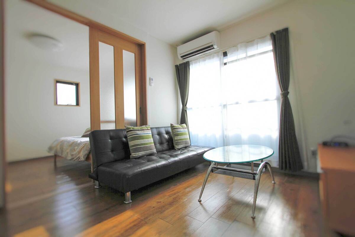 A modern living room with a black leather sofa, a glass coffee table, wooden flooring, a bed with a patterned bedspread, and a large window with curtains.