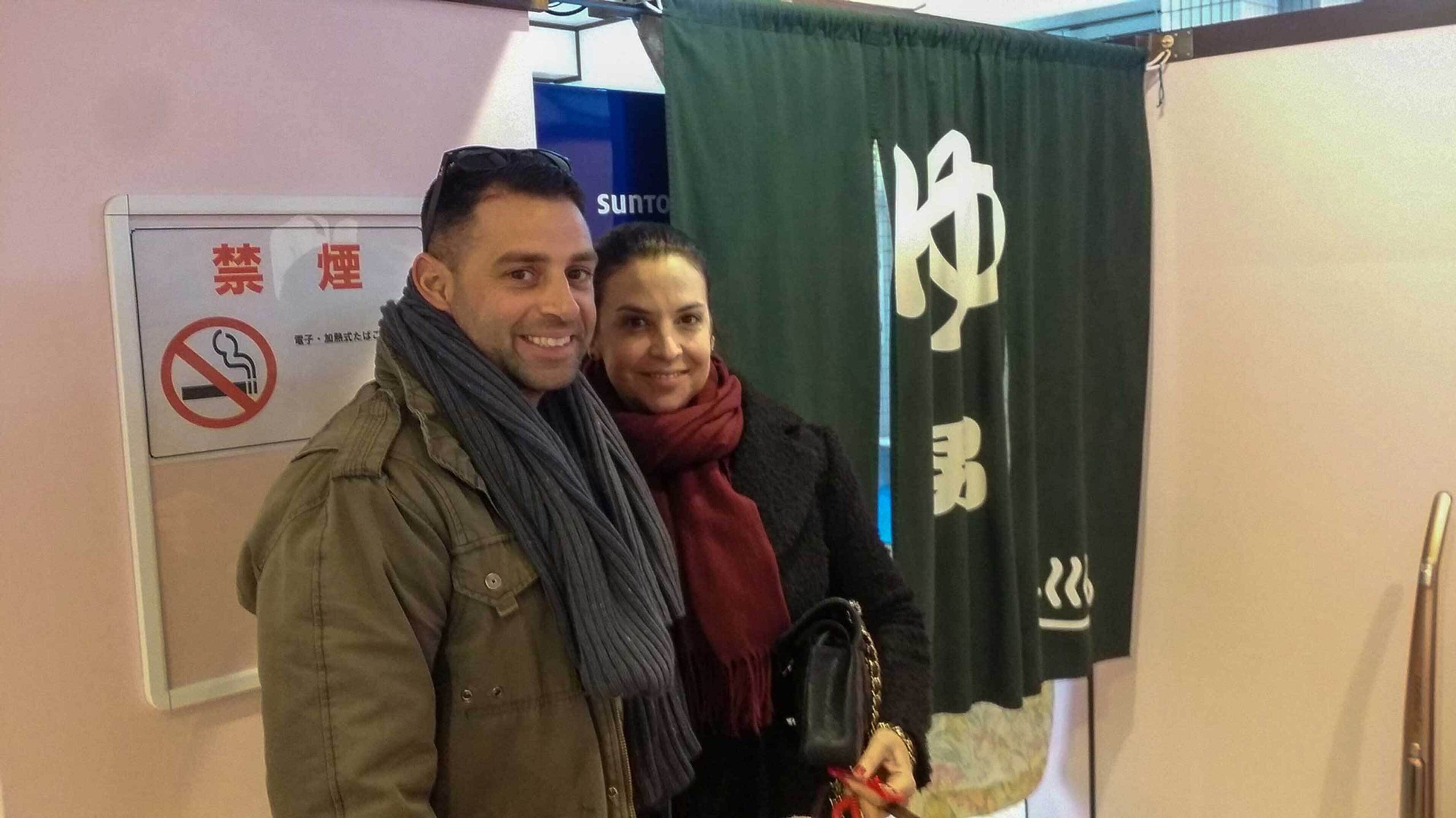 Due persone in piedi davanti a un cartello con il simbolo del divieto di fumare e una fascia verde con testo bianco.