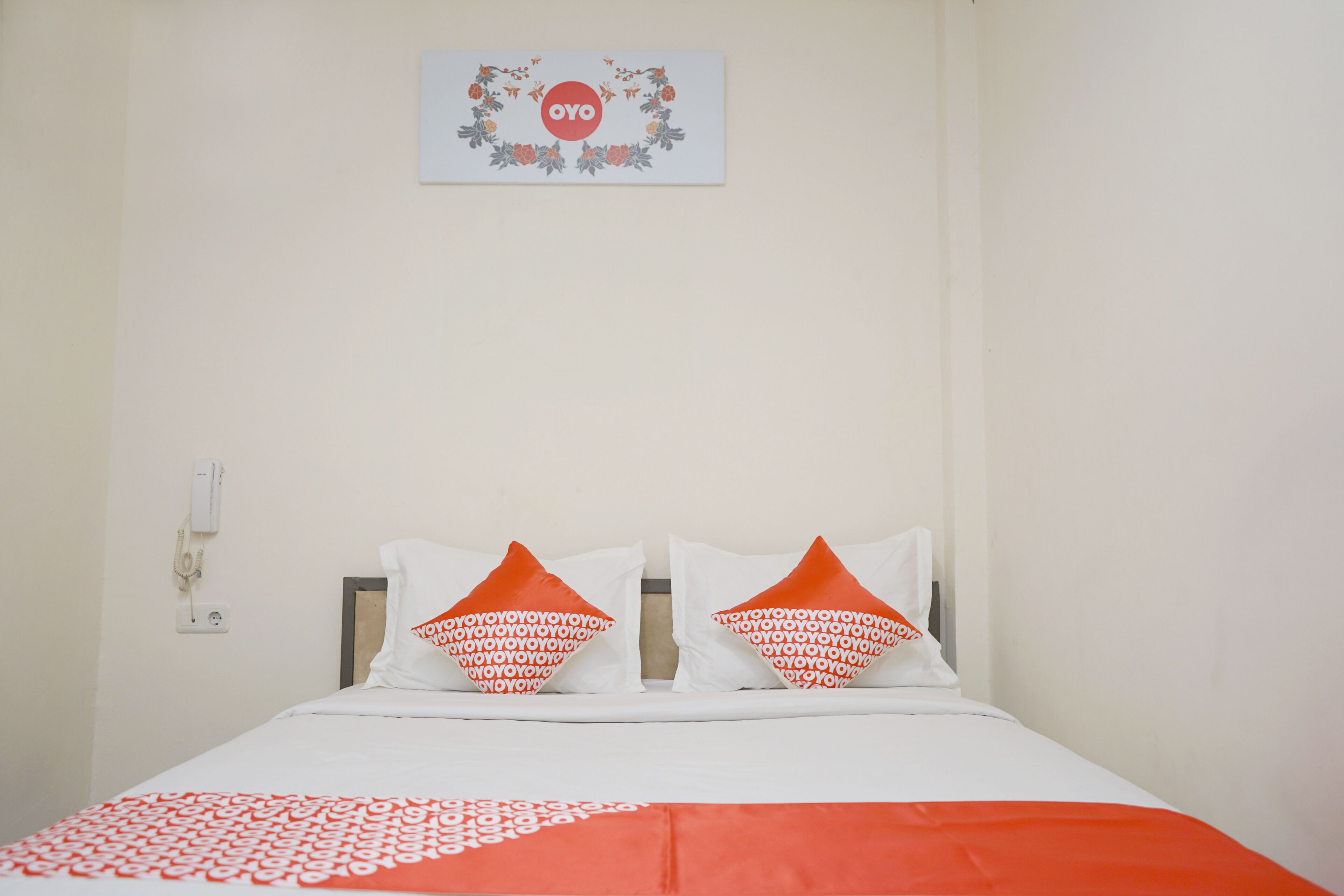 Habitación de hotel con cama, dos almohadas estampadas, una colcha, un cuadro en la pared y un enchufe.