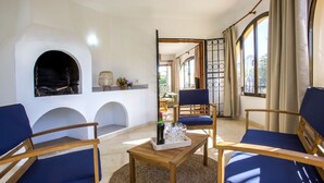 Une pièce avec une cheminée, deux fauteuils bleus, une table basse en bois et une vue par une fenêtre ornée de rideaux.