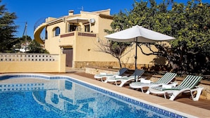 Un espace piscine avec des chaises longues et un parasol, un bâtiment doté d’un balcon incurvé, et un ciel d’un bleu limpide.