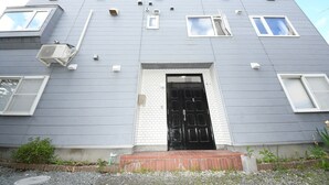 Un edificio de dos pisos con una puerta negra, ventanas con marco blanco y una unidad de aire acondicionado en la pared exterior.