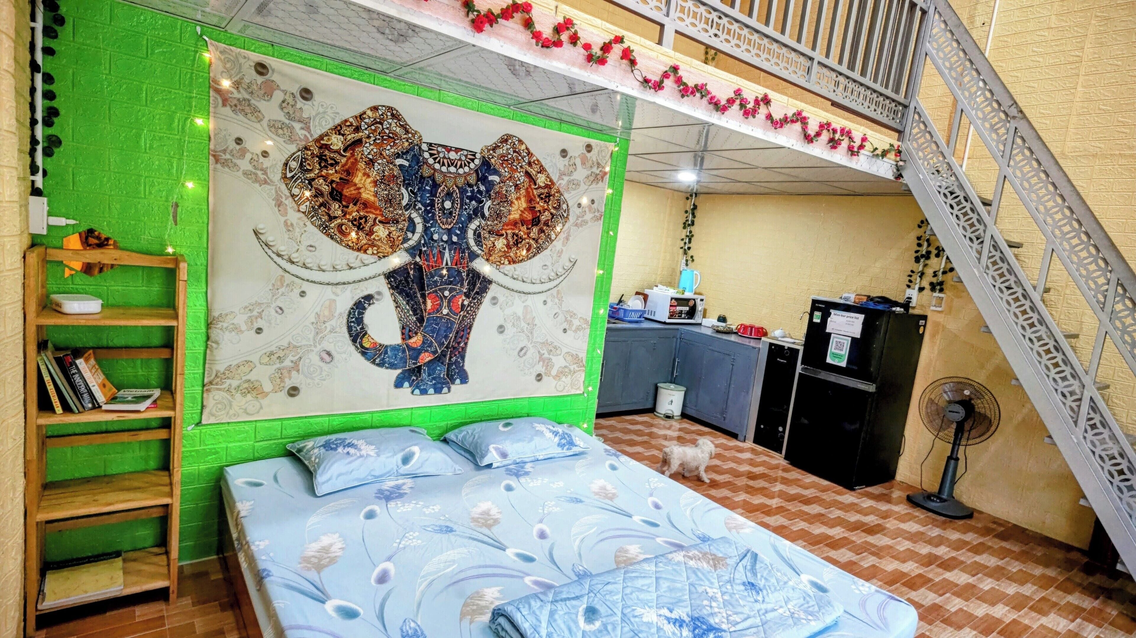 A bedroom with a large elephant mural, a bed with floral bedding, a wooden shelf with books, and a kitchenette area.