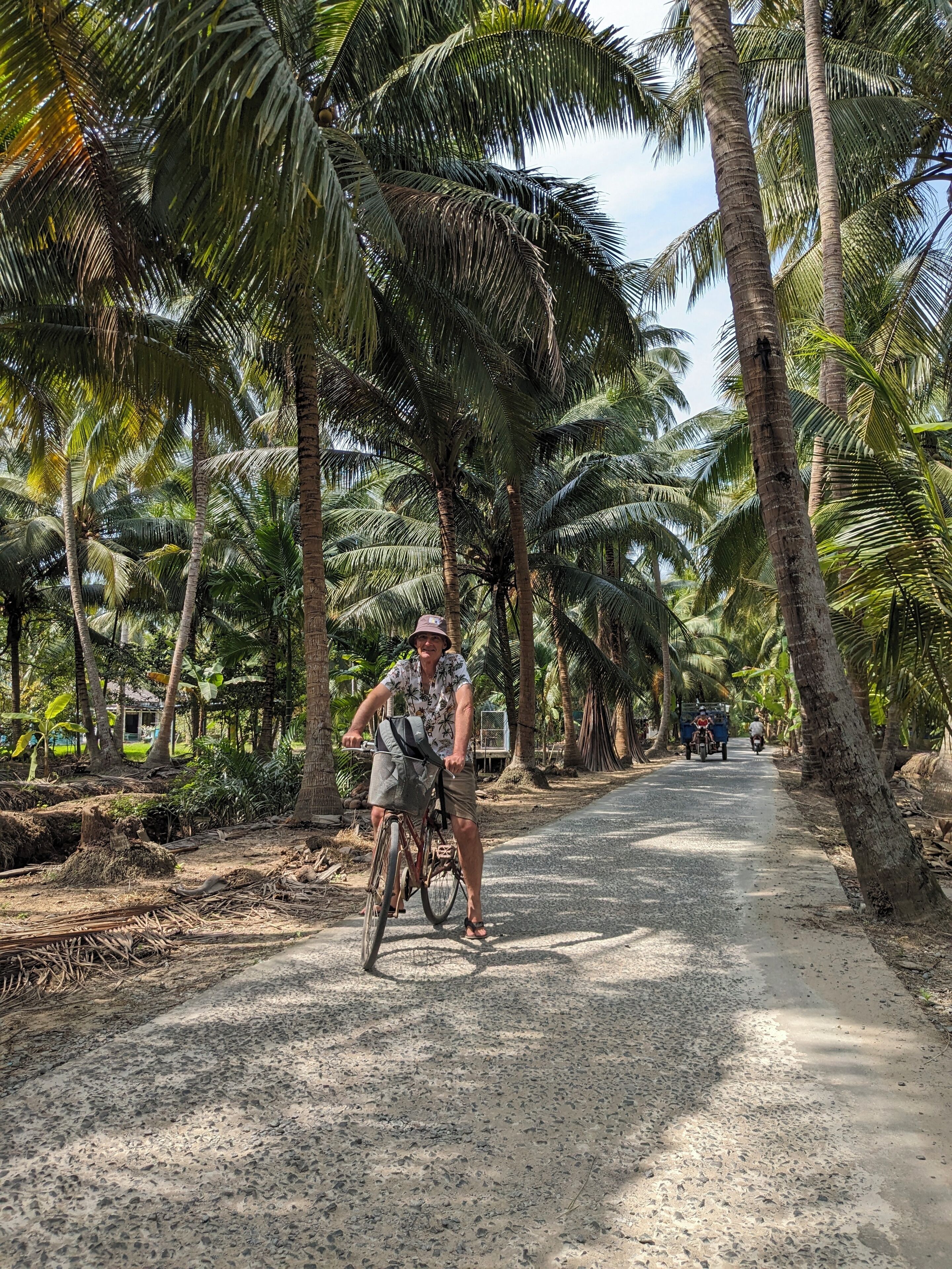 A paved road flanked by palm trees, with a cyclist and a motorbike in the distance.