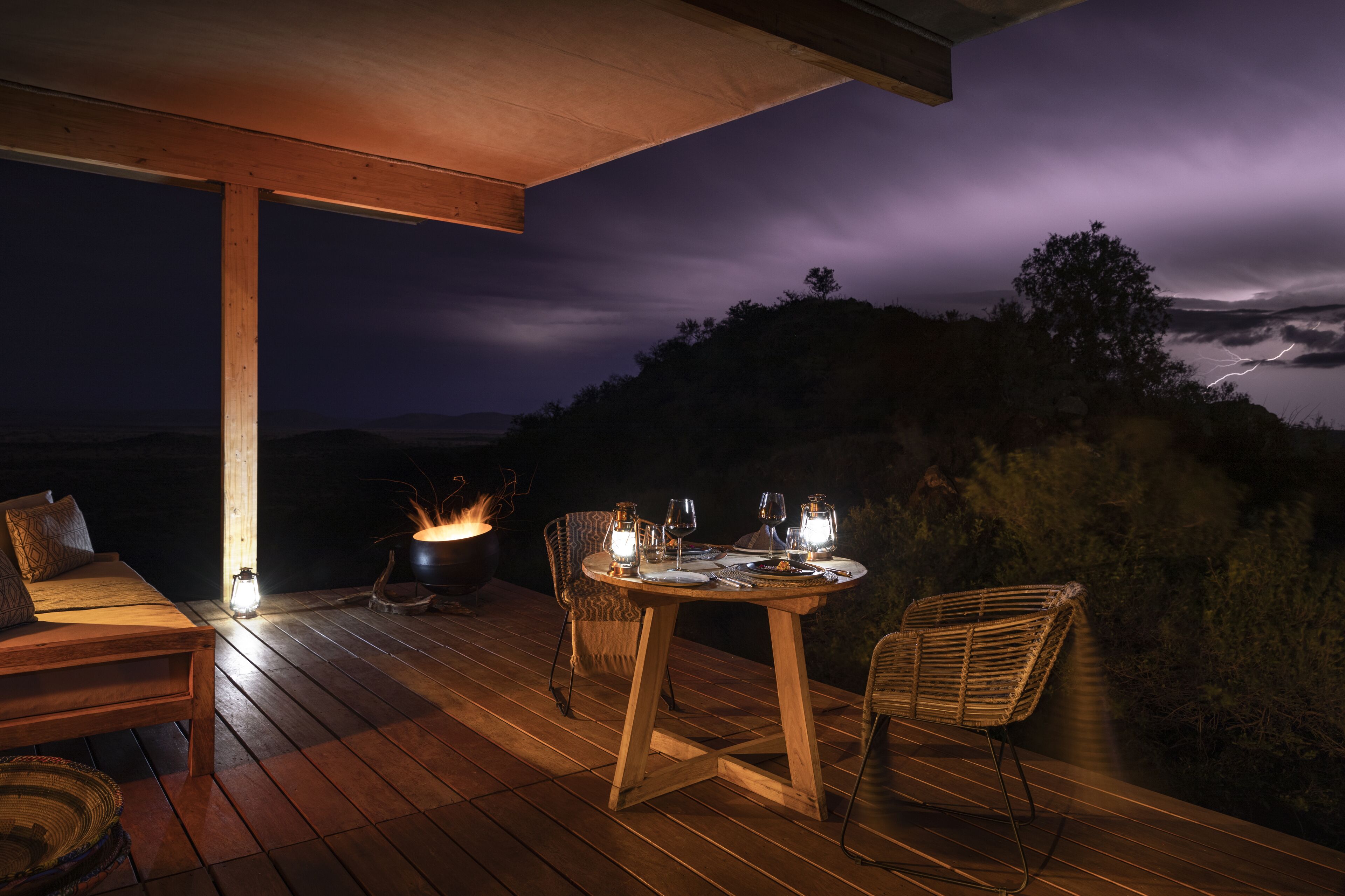 An outdoor dining area with a wooden table and chairs, a fire pit, and a view of the landscape.