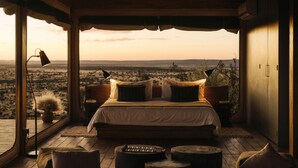 A bedroom with a large bed, a view of the desert, a lamp, and a potted plant. - Our Habitas Namibia (Windhoek)
