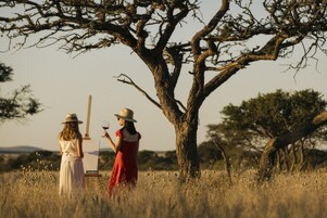 Dos mujeres, una vestida de rojo y otra de blanco, están de pie en un campo con un árbol y un caballete.