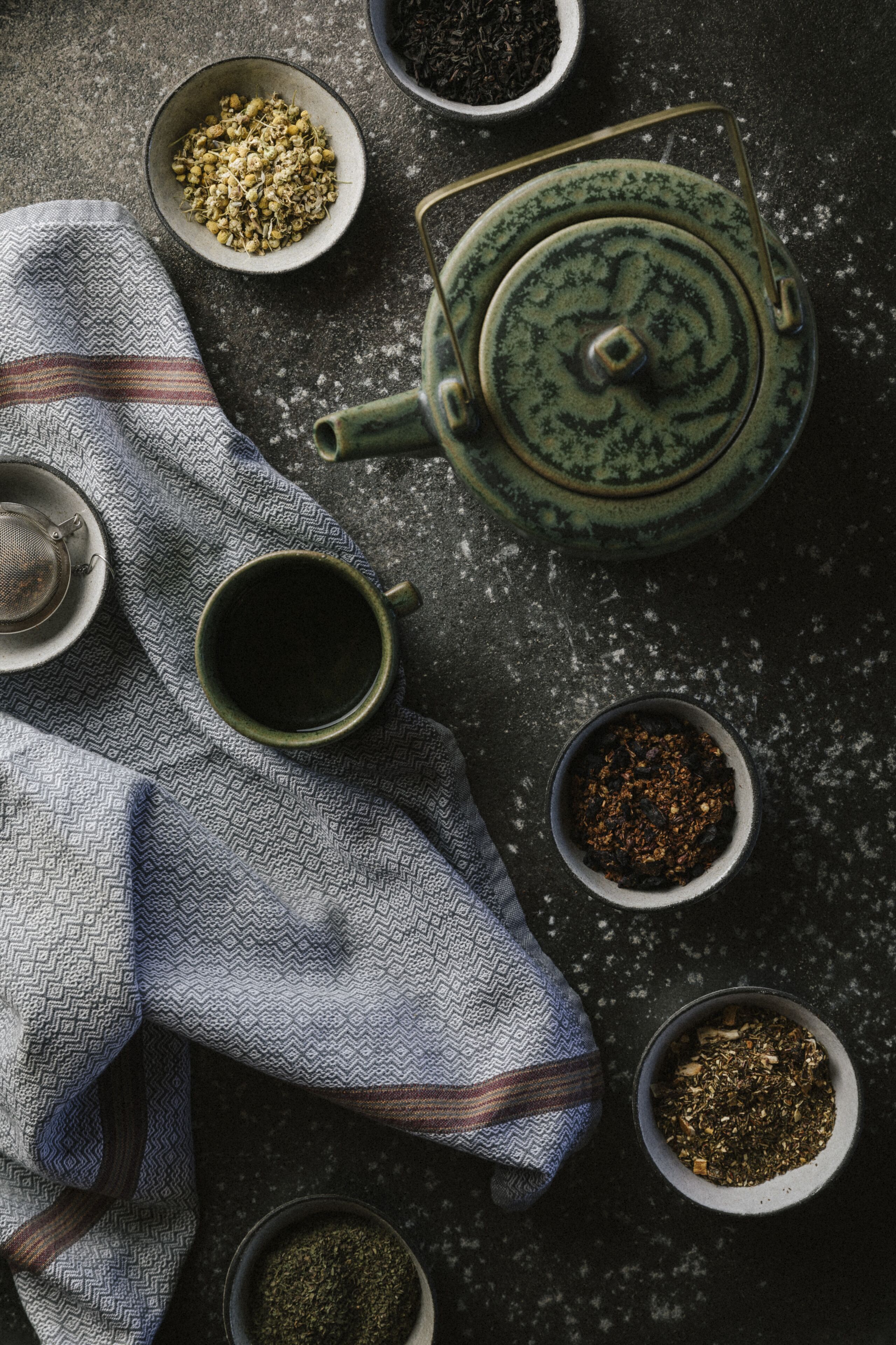 A top-down view of a tea set with a teapot, cups, and various loose tea leaves on a dark surface.