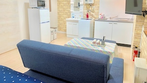 Una kitchenette compacta con microondas, refrigerador y un lavamanos pequeño. Hay un sofá y una mesa con mantel colorido.