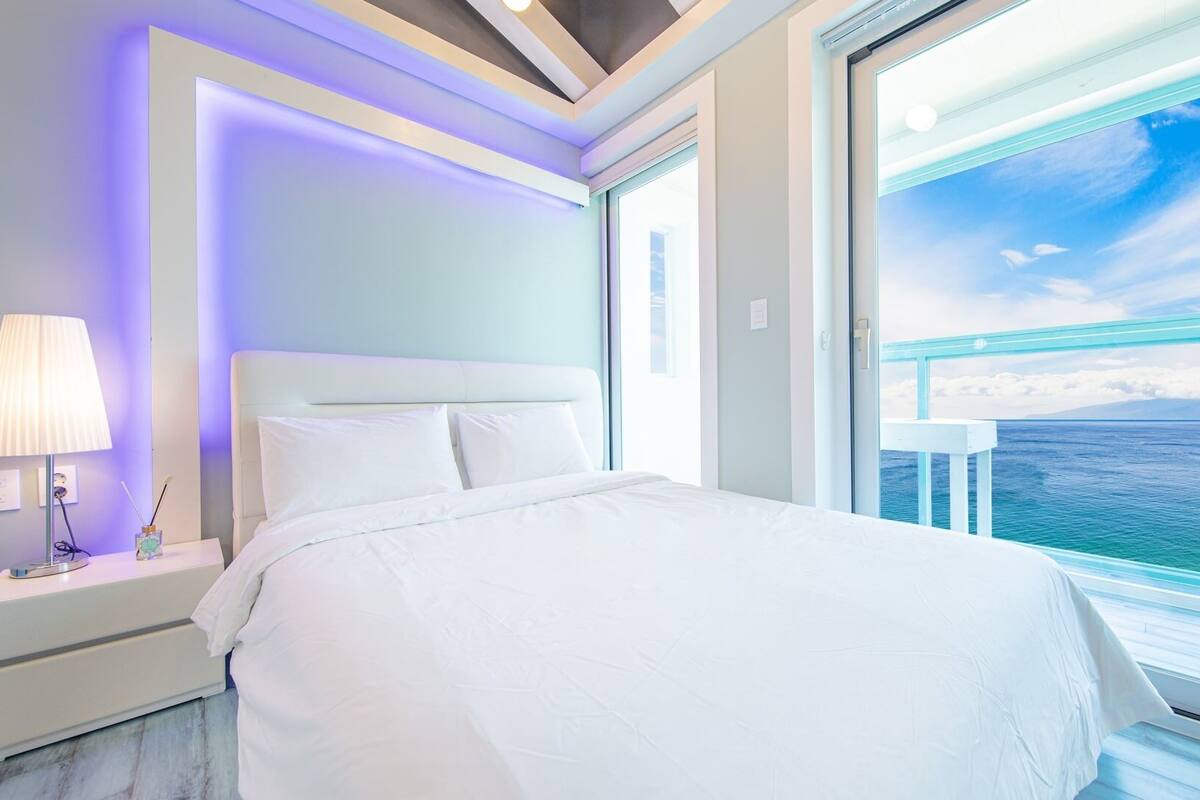 A bedroom with a bed, a nightstand, a lamp, and a view of the ocean through a balcony.